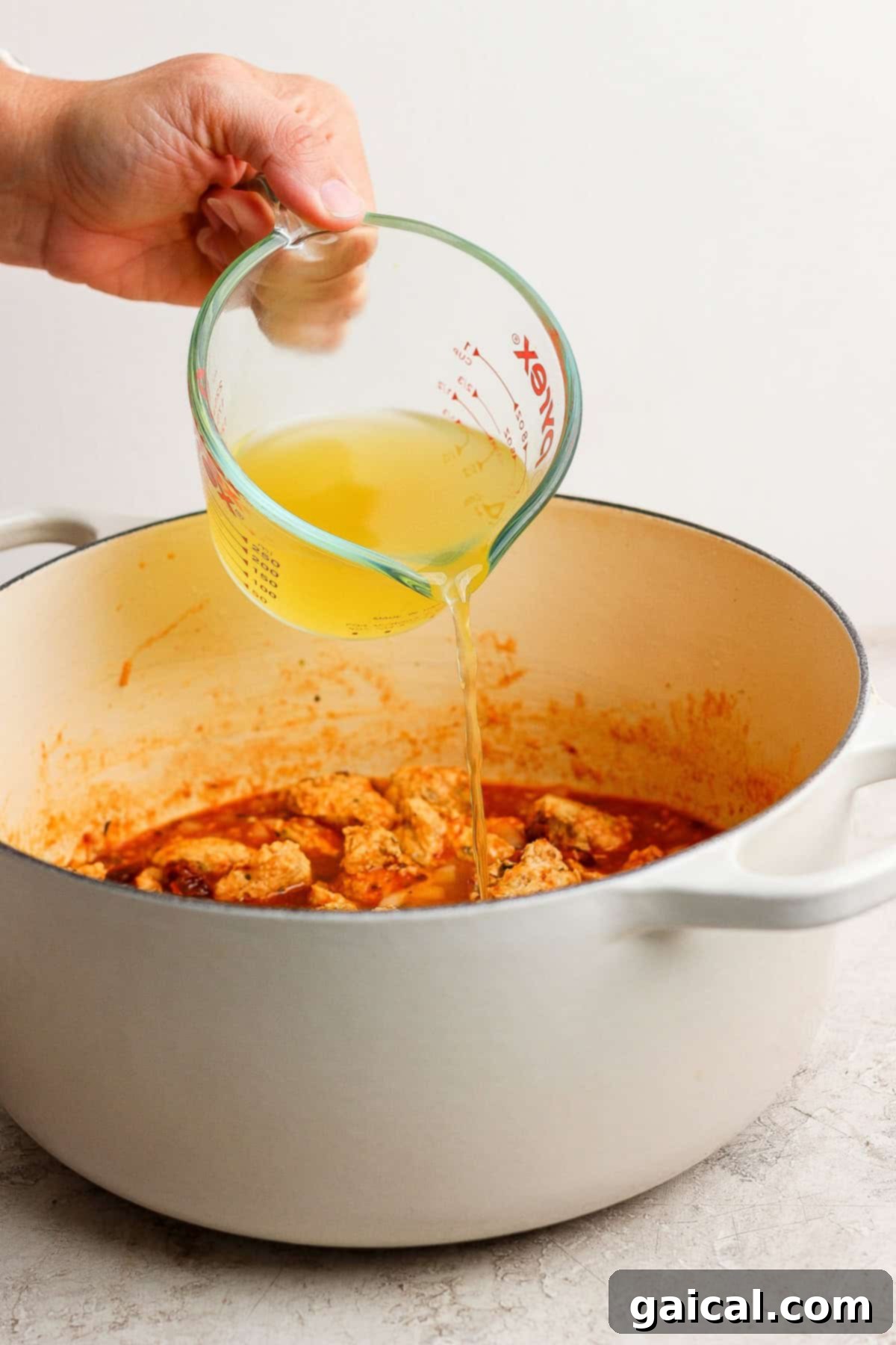 A stream of rich chicken broth being poured into a Dutch oven filled with simmering ingredients.