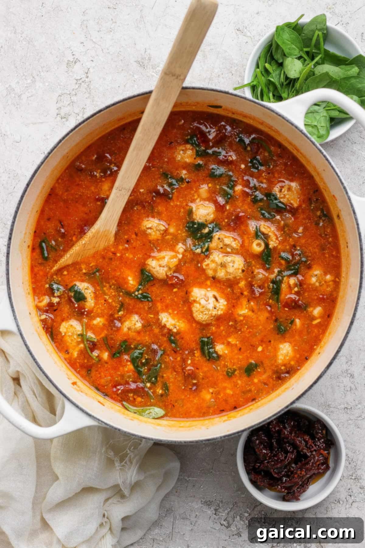 A large pot of Marry Me Chicken Soup, showcasing its rich tomato broth, tender chicken pieces, and vibrant spinach, ready to be served. Nearby are bowls of fresh ingredients including spinach and sun-dried tomatoes.