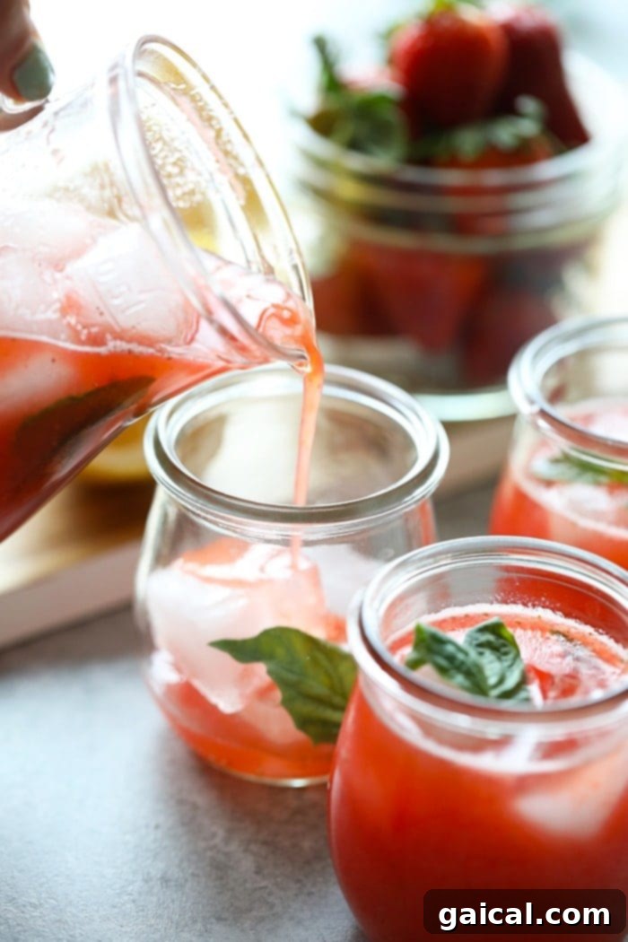 Berry Lemonade Bliss 7 Homemade strawberry basil lemonade being gracefully poured from a clear glass pitcher into a tall glass filled with ice, garnished with a fresh basil leaf.