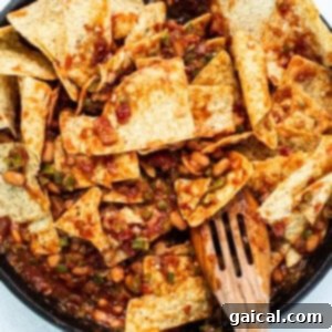 Mixing crispy tortilla chips into the rich chilaquiles sauce in a cast iron skillet, ensuring every chip is coated.