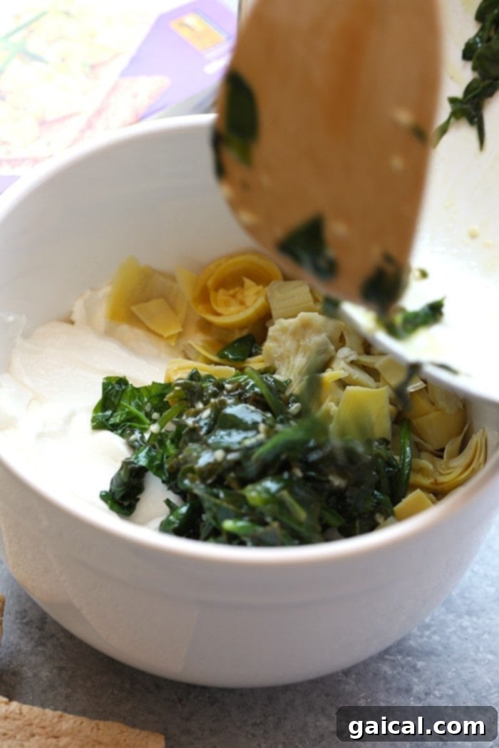 A vibrant collection of fresh ingredients for spinach artichoke dip, including green spinach leaves, canned artichoke hearts, softened cream cheese, and a bowl of Greek yogurt, artfully arranged on a kitchen counter.