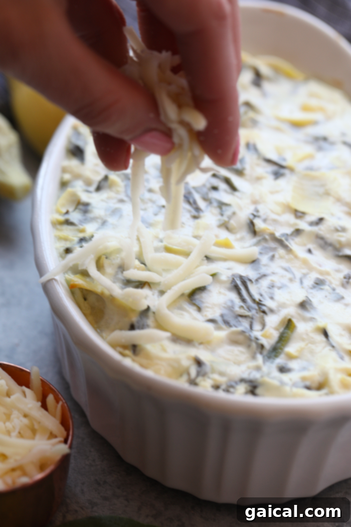Uncooked spinach artichoke dip mixture neatly arranged in a white baking dish, topped with a generous layer of shredded mozzarella cheese, glistening and ready for the oven.