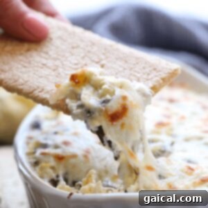 A person is dipping a cracker into a bowl of Lightened Up Spinach and Artichoke Dip, showcasing the dip's creamy, melted cheese texture.