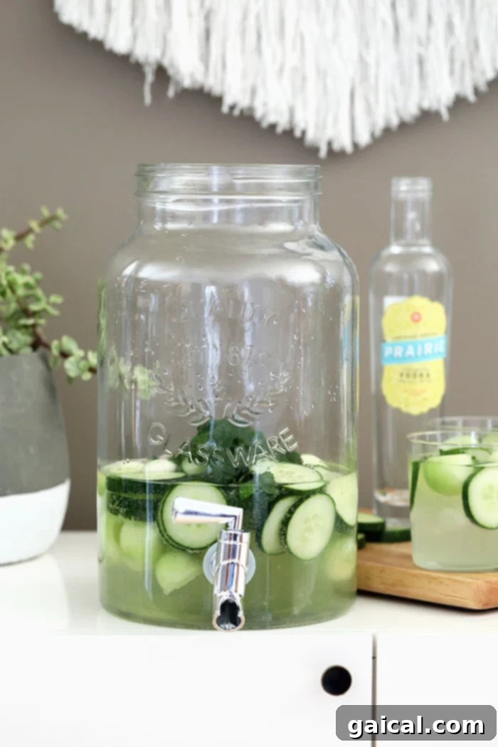 Cucumber Vodka Drink Dispenser for Parties A large, rustic-chic mason jar drink dispenser filled to the brim with a clear and inviting cucumber vodka cocktail. The drink is visibly infused with numerous fresh cucumber slices and honeydew melon balls, creating an appealing display that promises delicious refreshment. The dispenser is ready for serving at any party or casual gathering.