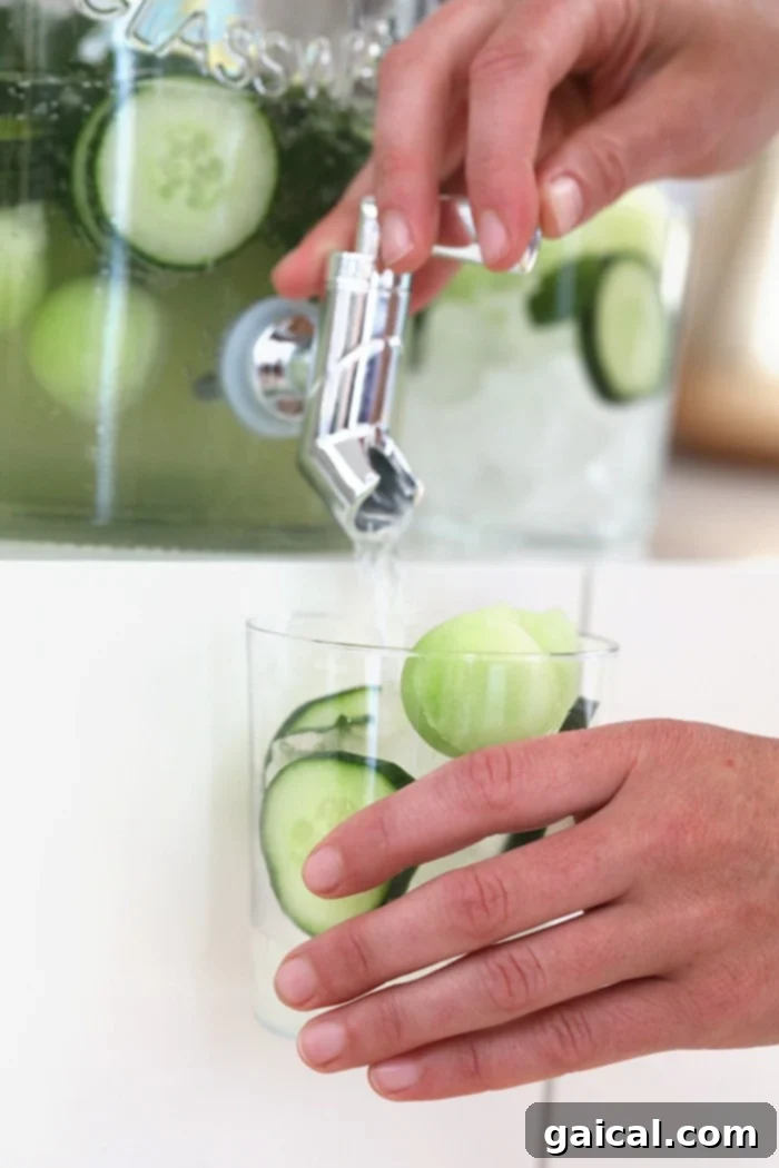 Serving Refreshing Cucumber Vodka Cocktails A hand is actively dispensing a sparkling cucumber vodka cocktail from a sleek glass drink dispenser into individual glasses. The glasses are ready to be served, each featuring a refreshing garnish of cucumber slices and honeydew melon balls, highlighting the convenience and visual appeal of this large batch drink.