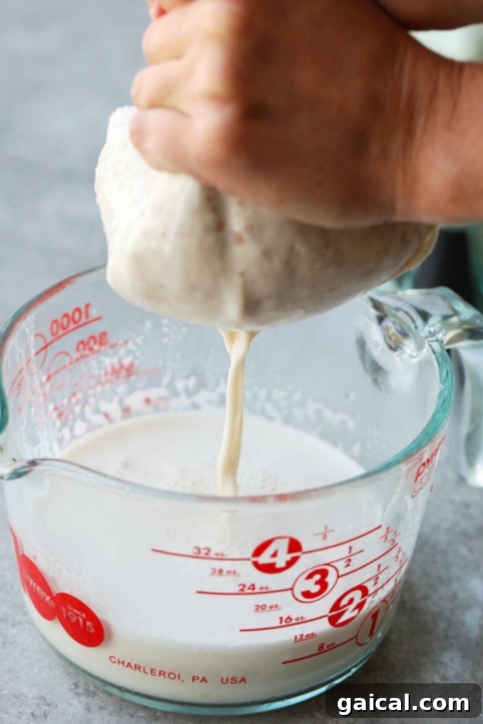 The action of straining freshly blended almond milk through a nut milk bag into a glass pitcher, creating smooth, pulp-free milk.