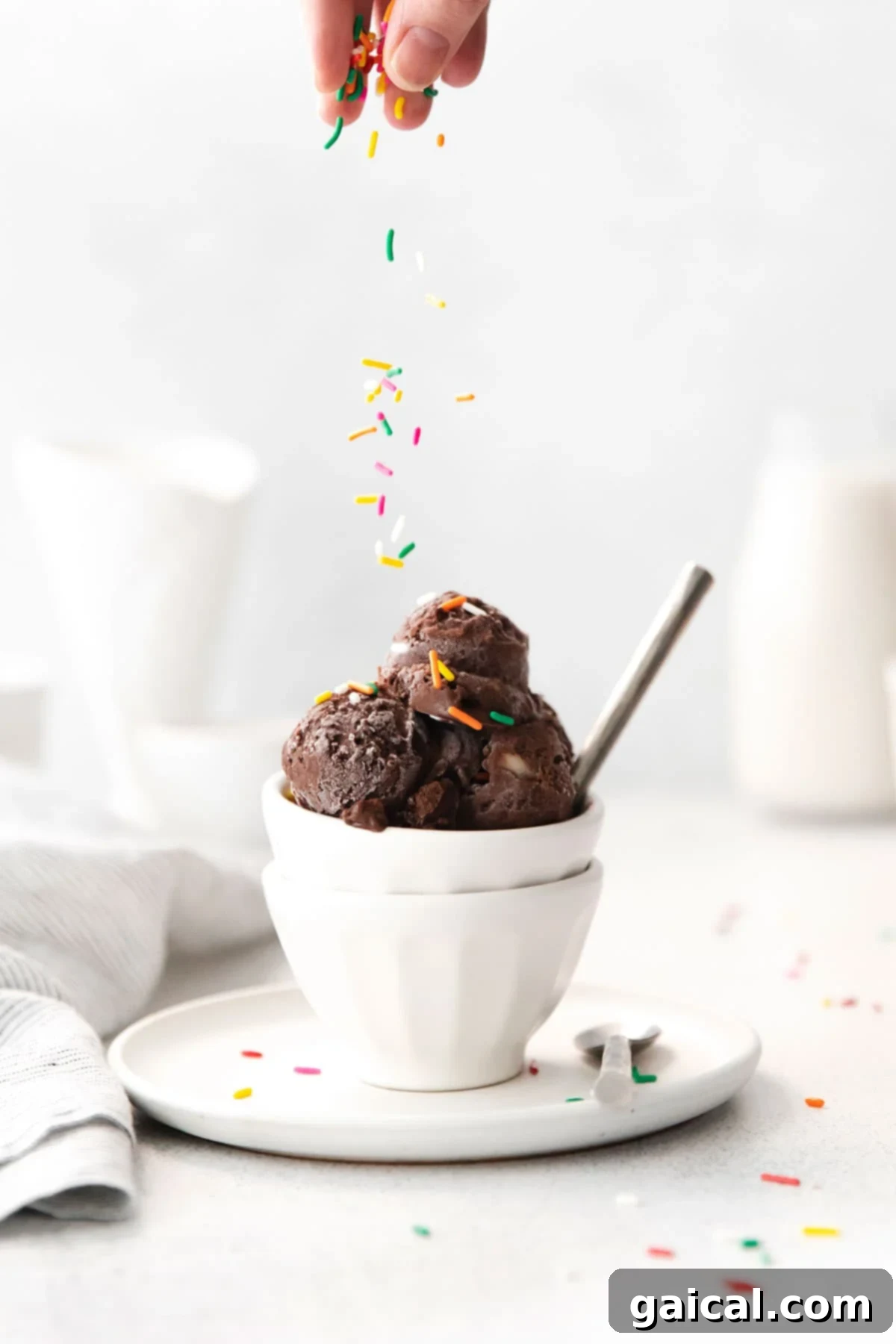 This chocolate protein ice cream recipe is dairy-free and made with frozen bananas, cocoa powder, chocolate protein powder, and almond milk! With 15 grams of protein per serving, it is a healthy treat too! A close-up shot of a hand gracefully sprinkling a generous amount of colorful rainbow sprinkles over a creamy portion of chocolate protein ice cream in a pristine white bowl, showcasing the final delicious topping.