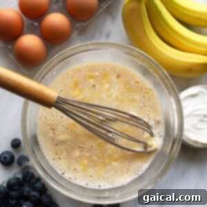 A large mixing bowl containing whisked eggs, honey, Greek yogurt, mashed banana, almond milk, and vanilla extract, forming a creamy wet mixture.