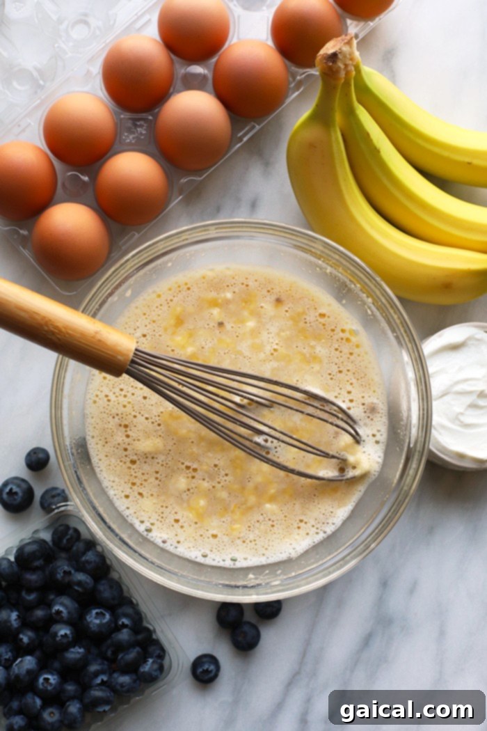 These healthy blueberry muffins get their natural sweetness from honey and bananas, and their incredible moisture from Greek yogurt. A perfect pairing with your morning coffee! A glass mixing bowl filled with creamy wet ingredients for muffins, including mashed banana, honey, and Greek yogurt, ready to be combined with dry ingredients.