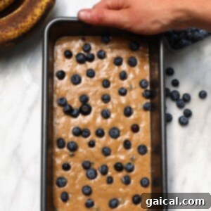 A person holding a loaf pan filled with blueberry banana bread, freshly baked and ready to cool, with a vibrant blueberry topping.
