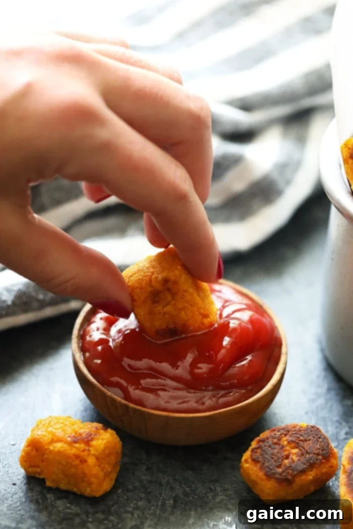 These healthy sweet potato tater tots are easy to make, delicious, and something the whole family will love! A hand dipping a crispy sweet potato tater tot into a vibrant red ketchup, illustrating a classic serving suggestion.