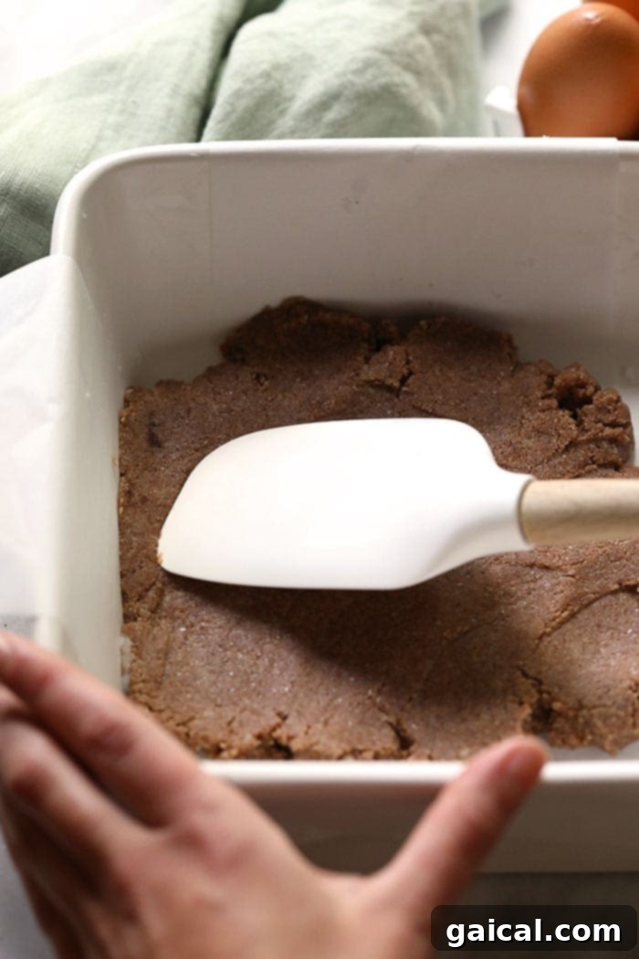 Wholesome Pumpkin Spice Bars 3 A pair of hands using a spatula to evenly spread the almond flour pumpkin pie crust mixture across the bottom of a baking pan.