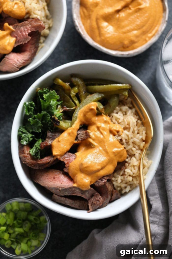 A beautifully arranged meal prep bowl with sliced steak, roasted peppers and onions, brown rice, and a side of green kale salad.