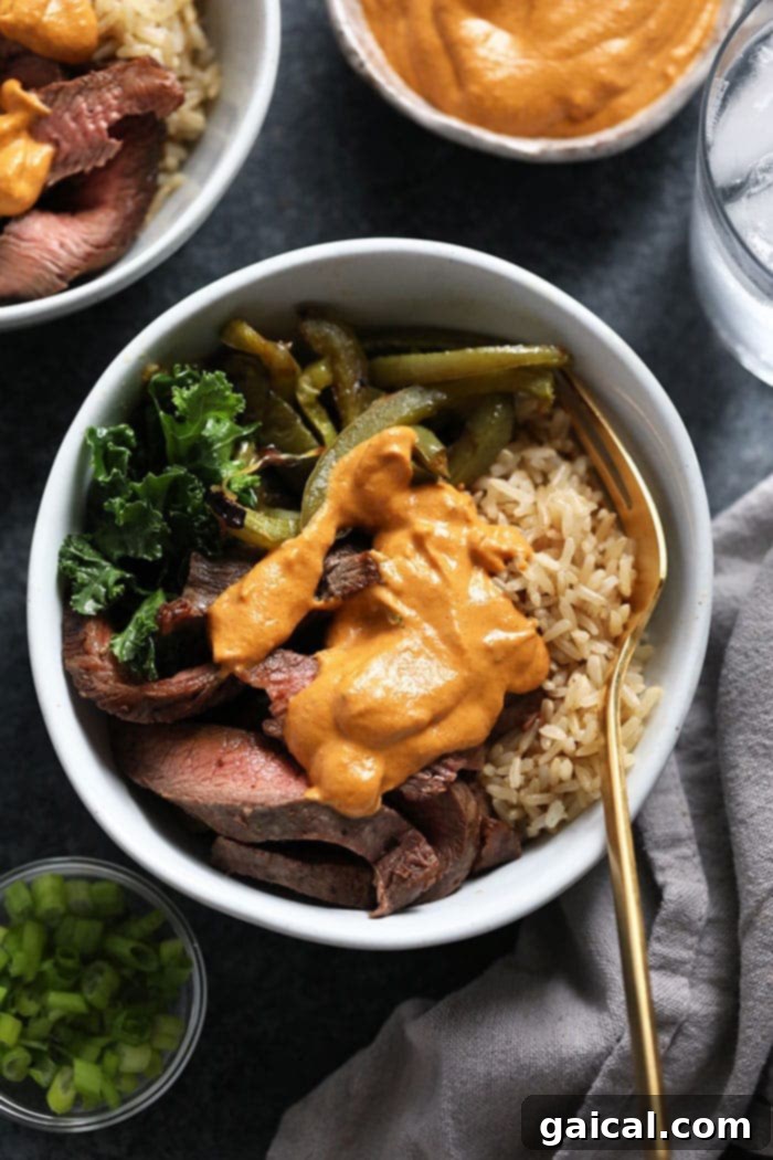 A close-up of an assembled meal prep bowl with all the components clearly visible: steak, peppers, onions, rice, kale, and vegan queso.
