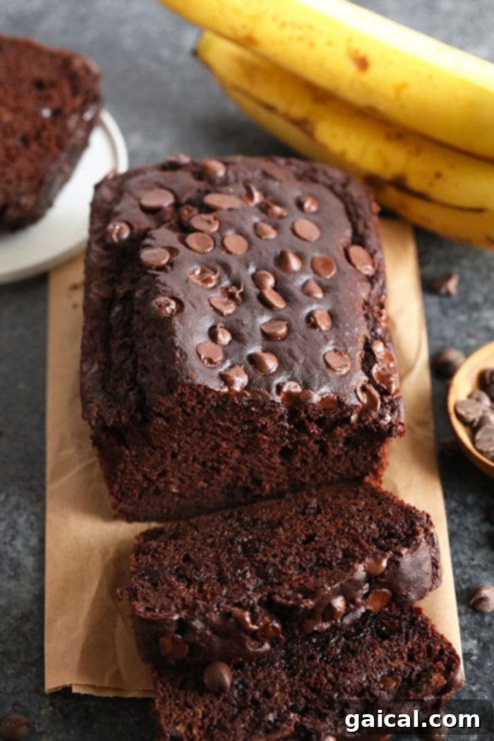 A perfectly sliced chocolate chocolate chip banana bread on a cutting board, with whole bananas in the background.