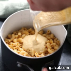A person is pouring creamy vegan cheese sauce over cooked pasta in a pan, ready to mix.