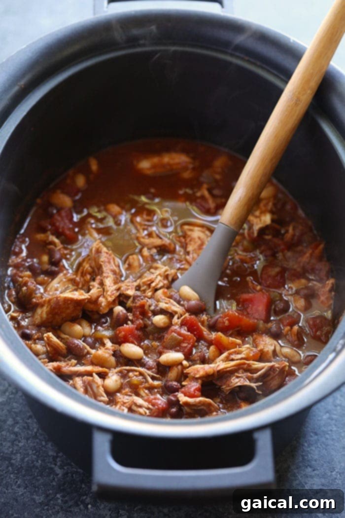 Comforting Chicken Chili: Perfect for a cozy meal. Generous serving of crockpot chicken chili in a rustic ceramic bowl, topped with a dollop of sour cream and fresh green onions, on a wooden surface.