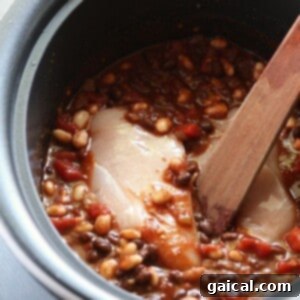 Effortless Slow Cooker Chicken Chili 3 A crockpot full of chili ingredients including beans, tomatoes, and chicken breasts, with a wooden spoon for mixing.