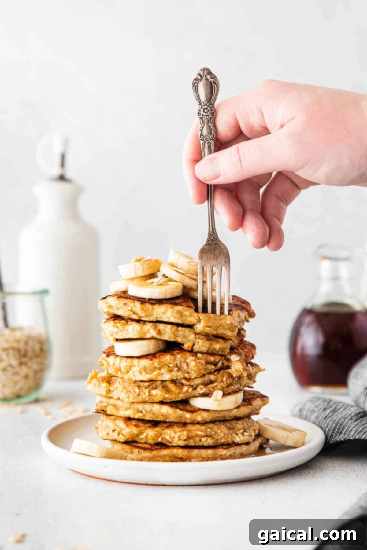 Fork biting into a stack of fluffy banana oatmeal pancakes, drizzled with syrup and topped with fresh banana slices.
