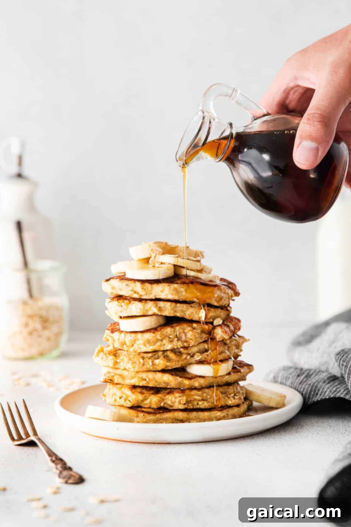 A stack of golden-brown banana oatmeal pancakes being generously drizzled with warm maple syrup.