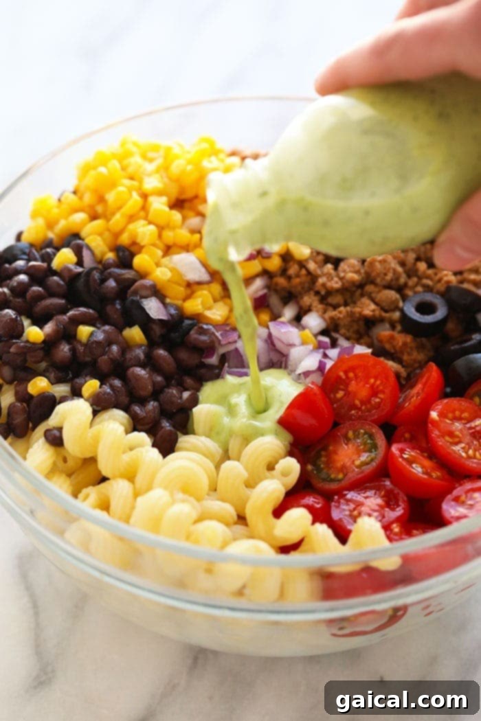 Delicious Mexican pasta salad that will serve a crowd. Bring this taco pasta salad to your next BBQ or make it for meal prep! A stream of creamy cilantro lime dressing being poured over a large bowl of taco pasta salad ingredients