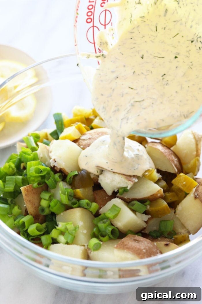 Thick, creamy vegan dressing being poured generously over cooked potatoes and fresh herbs in a mixing bowl.