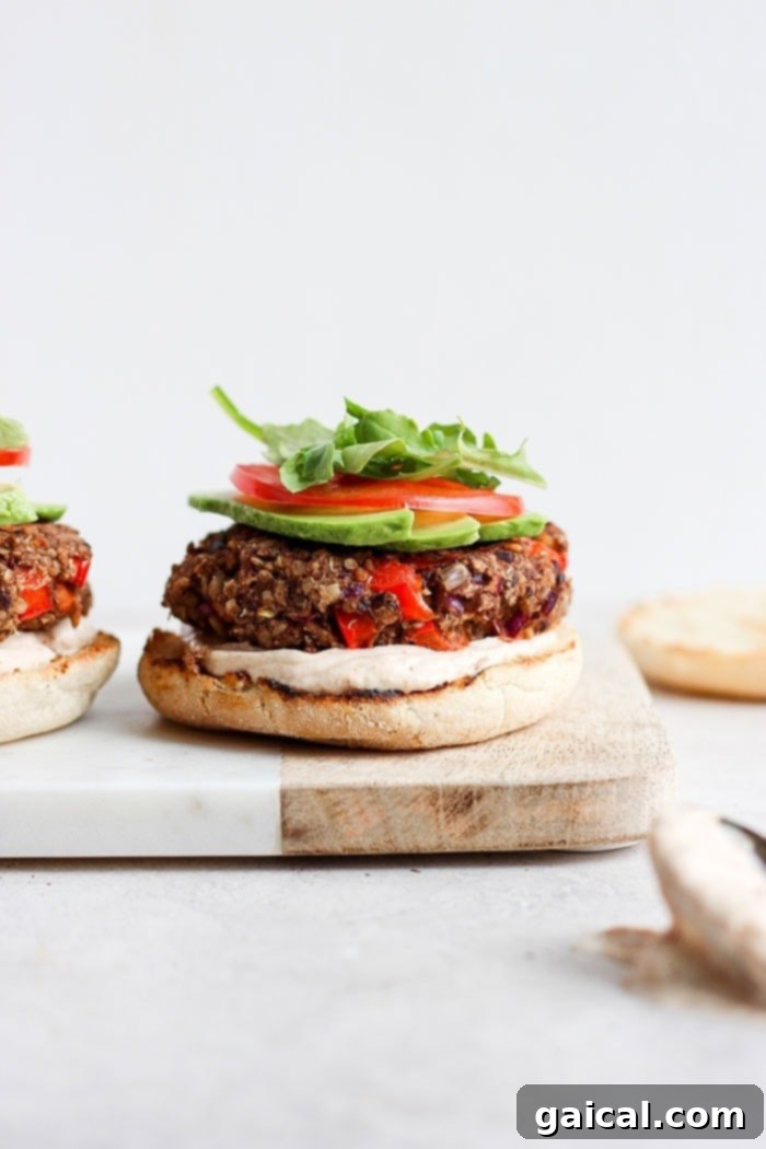 A black bean quinoa burger on an English muffin, beautifully garnished with sauce, avocado, tomato, and arugula, on a cutting board.