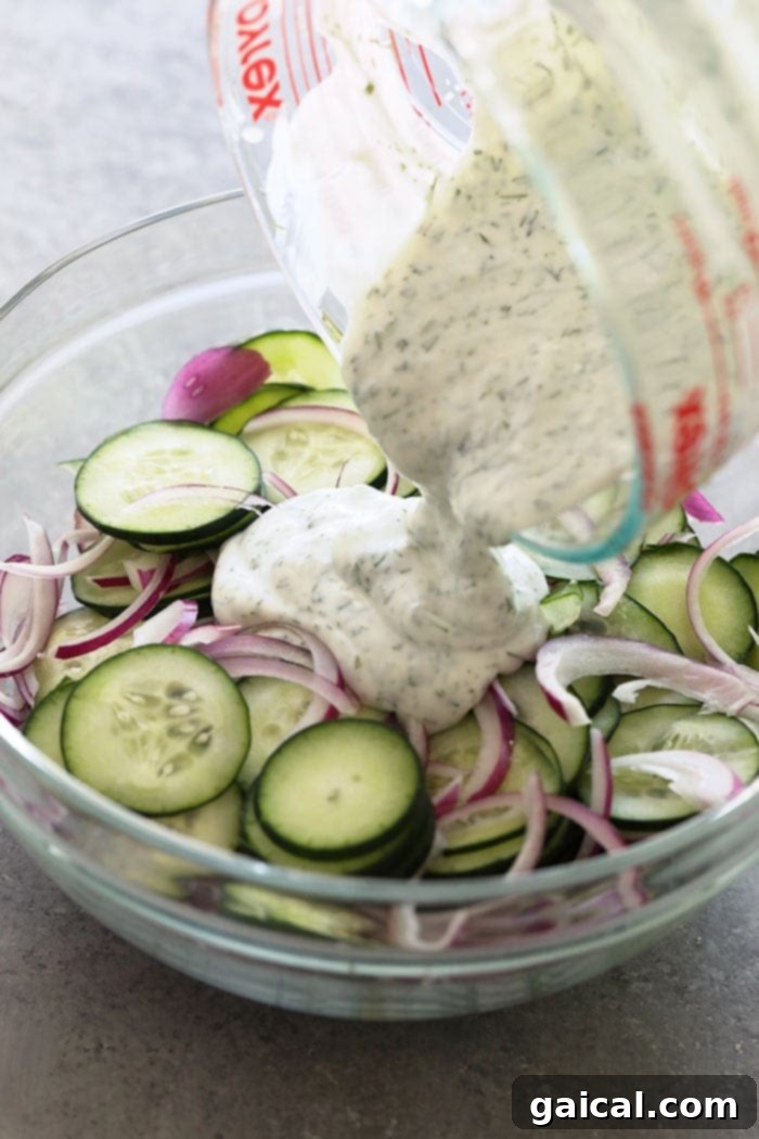 Toss thinly sliced cucumbers and red onion in a rich, creamy Greek yogurt dressing for the perfect texture and flavor. Pouring creamy dressing over sliced cucumbers and red onion in a bowl.