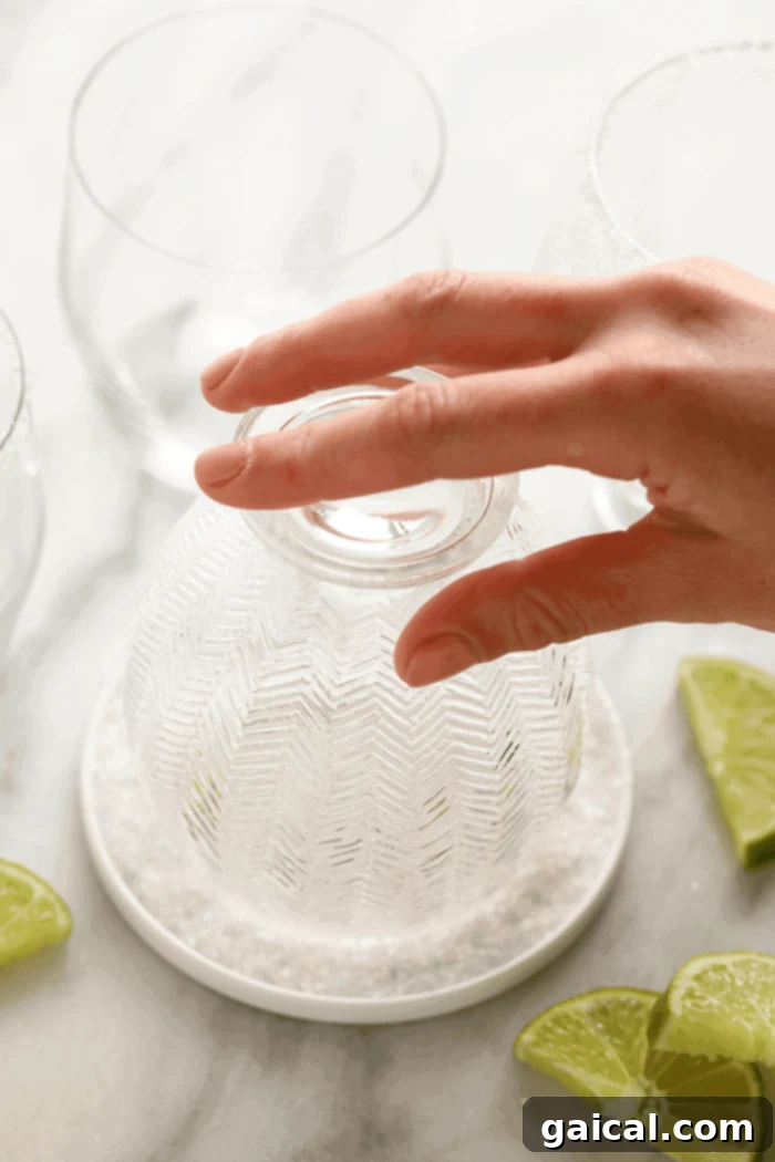 Master the classic margarita glass rimming technique with just water and coarse salt. A hand dipping the rim of a margarita glass into coarse sea salt spread on a plate, preparing it for serving.