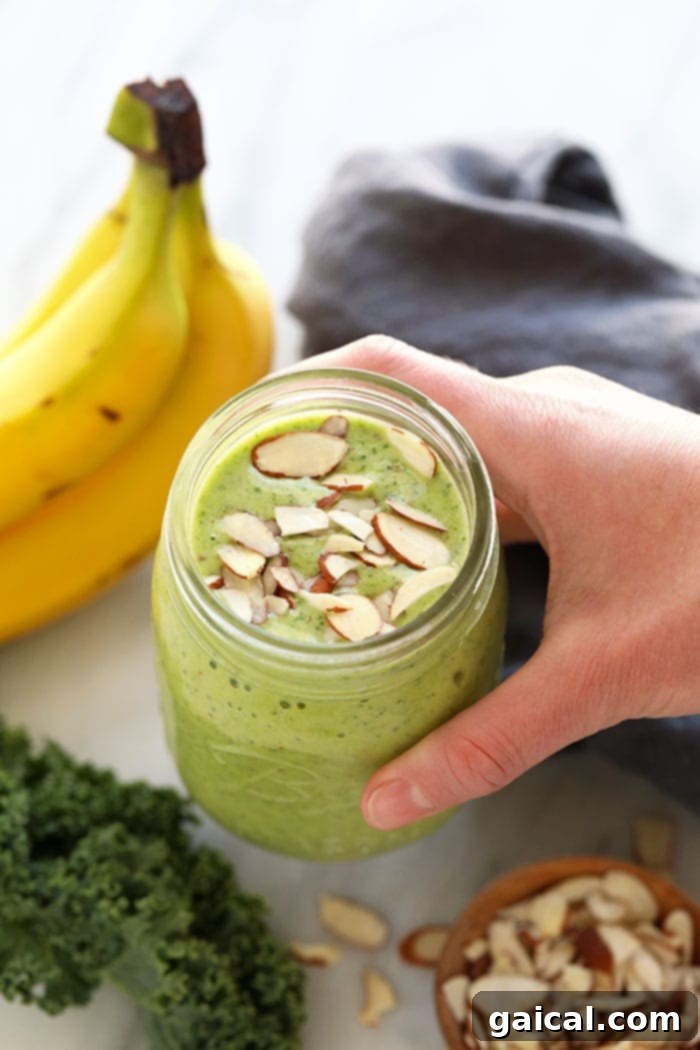 Get your daily greens here! This is the most delicious kale smoothie you'll ever make! It's packed with nutrient dense kale, frozen bananas and the perfect flavor punch of orange juice and fresh ginger. Whip up this green smoothie in 5 minutes and you're off and running with a healthy breakfast or snack on the go. A finished kale smoothie in a mason jar, elegantly topped with slivered almonds and a mint leaf.