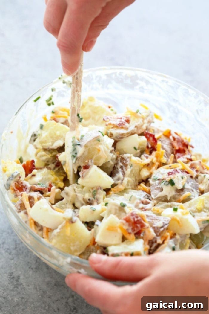 A hand mixing loaded baked potato salad in a large bowl, showcasing the rich texture and ingredients.
