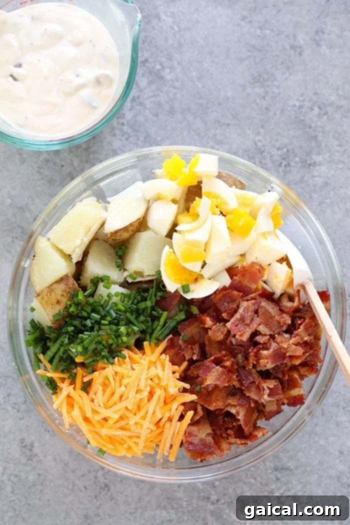 Ingredients for loaded potato salad laid out on a kitchen counter: cooked potatoes, bacon, hard-boiled eggs, cheese, chives, and sauce components.
