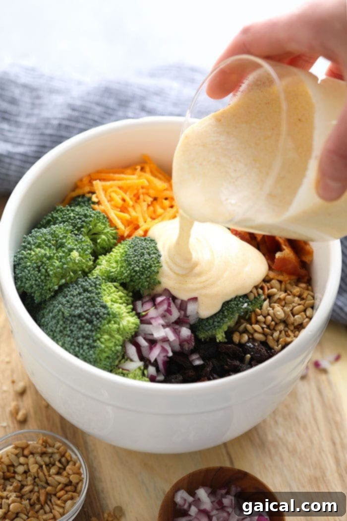 Pouring Dressing over Broccoli Salad Ingredients Creamy dressing being poured over a large bowl filled with healthy broccoli salad ingredients, including fresh broccoli, red onion, sunflower seeds, raisins, and cheddar cheese, before mixing.