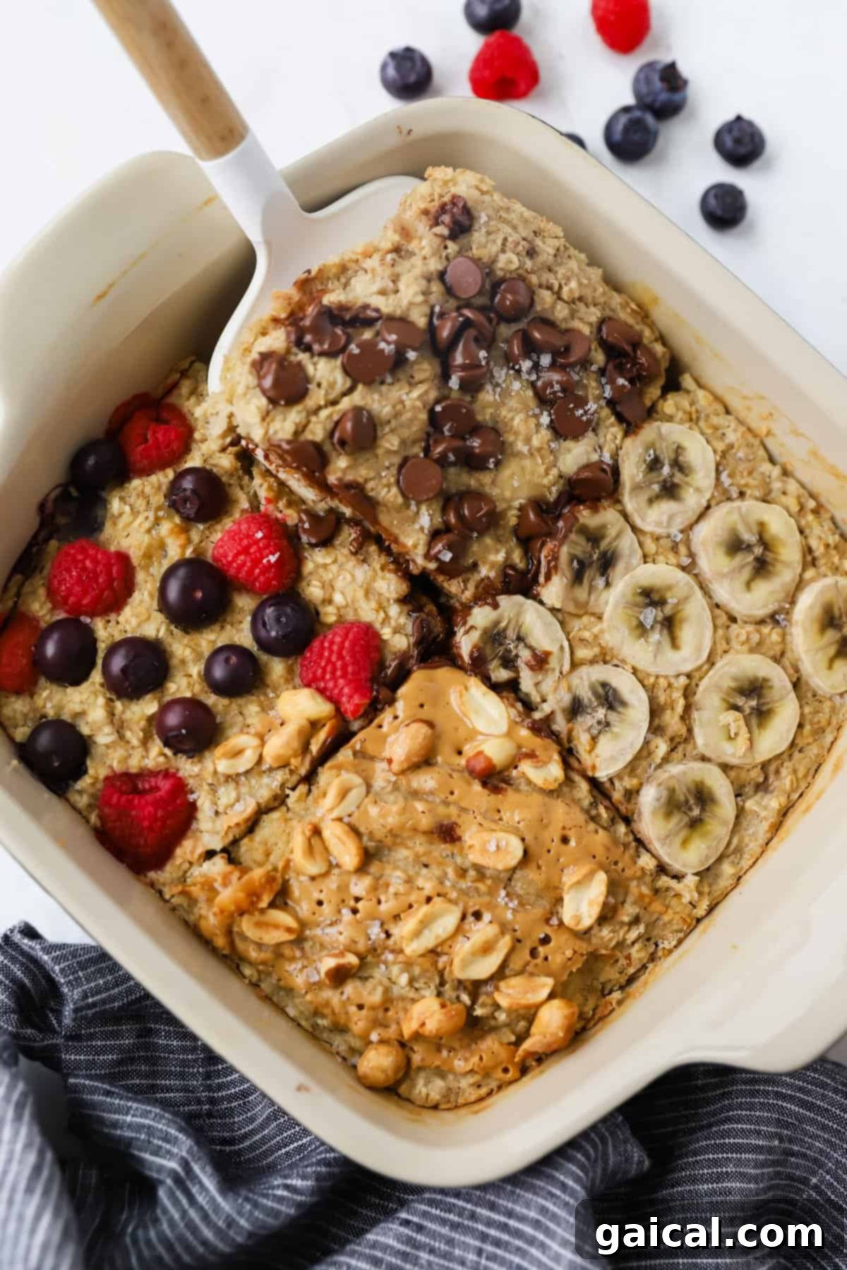 A baking dish with four sections of baked oatmeal topped with chocolate chips, banana slices, mixed berries, and peanut butter with peanuts. A spatula rests on one section.