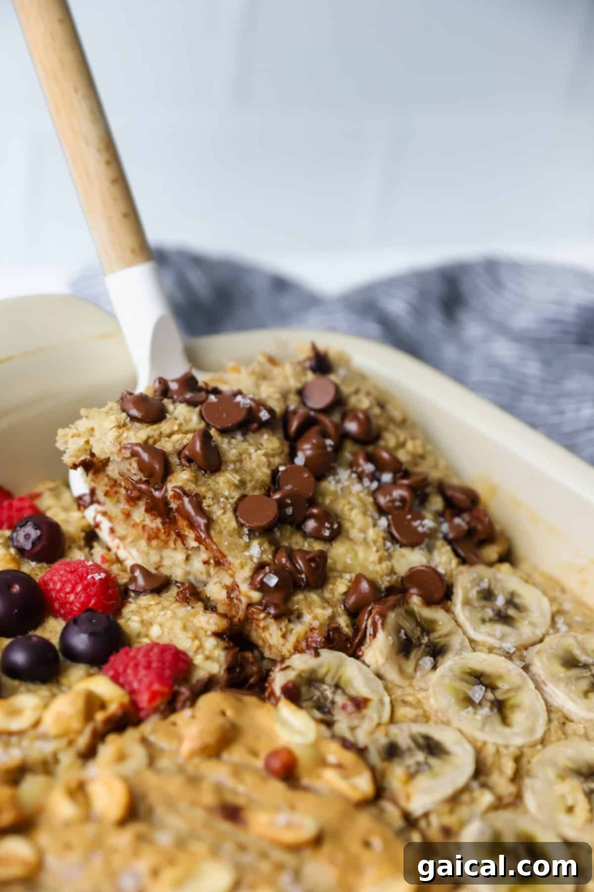A delicious baked oatmeal, bursting with chocolate chips, sliced bananas, berries, and nuts. A spatula gracefully lifts a portion of this comforting dish.