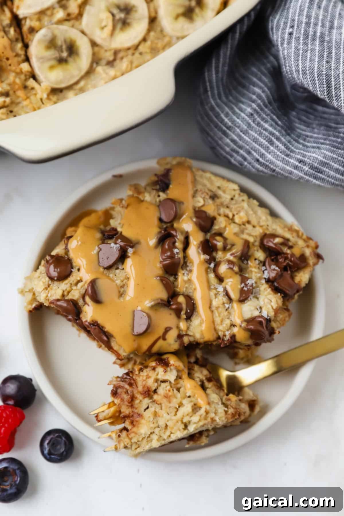 A plate with a slice of banana baked oatmeal, freshly baked, topped with chocolate chips and a peanut butter drizzle. A fork rests on the plate, accompanied by vibrant blueberries and a raspberry on the side.