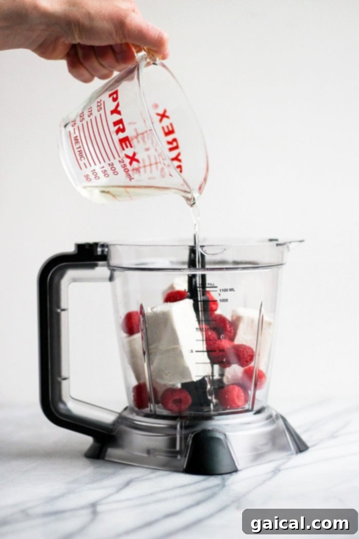 These raspberry cheesecake fat bombs are a delicious keto friendly dessert that tastes just like raspberry cheesecake! Make a big batch of these fat bombs and keep them in the freezer for an easy dessert all week long! Pouring melted coconut oil into a food processor with other ingredients, a crucial step in preparing keto fat bombs.