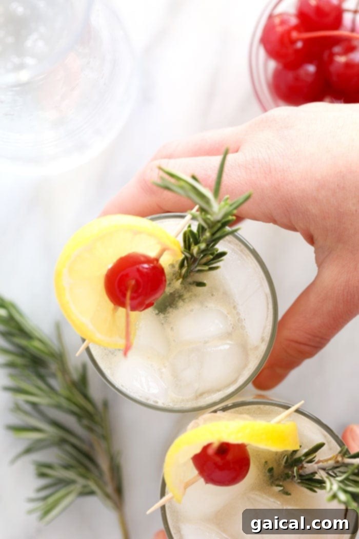 Over 15 Incredible Vodka Cocktail Recipes 13 A person holding a beautifully garnished glass of Rosemary Vodka Collins with cherries and a sprig of fresh rosemary.