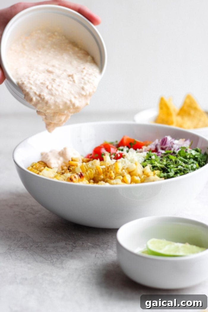 This Mexican Street Corn Salad, featuring a delicious Greek yogurt sauce, is the perfect side dish for any meal. A hand pouring the creamy white sauce over a bowl of vibrant Mexican Street Corn Salad.