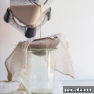 Craft Your Own Oat Milk 6 A clean tea towel being carefully placed over a fine-mesh sieve and a pitcher, preparing to strain homemade oat milk.