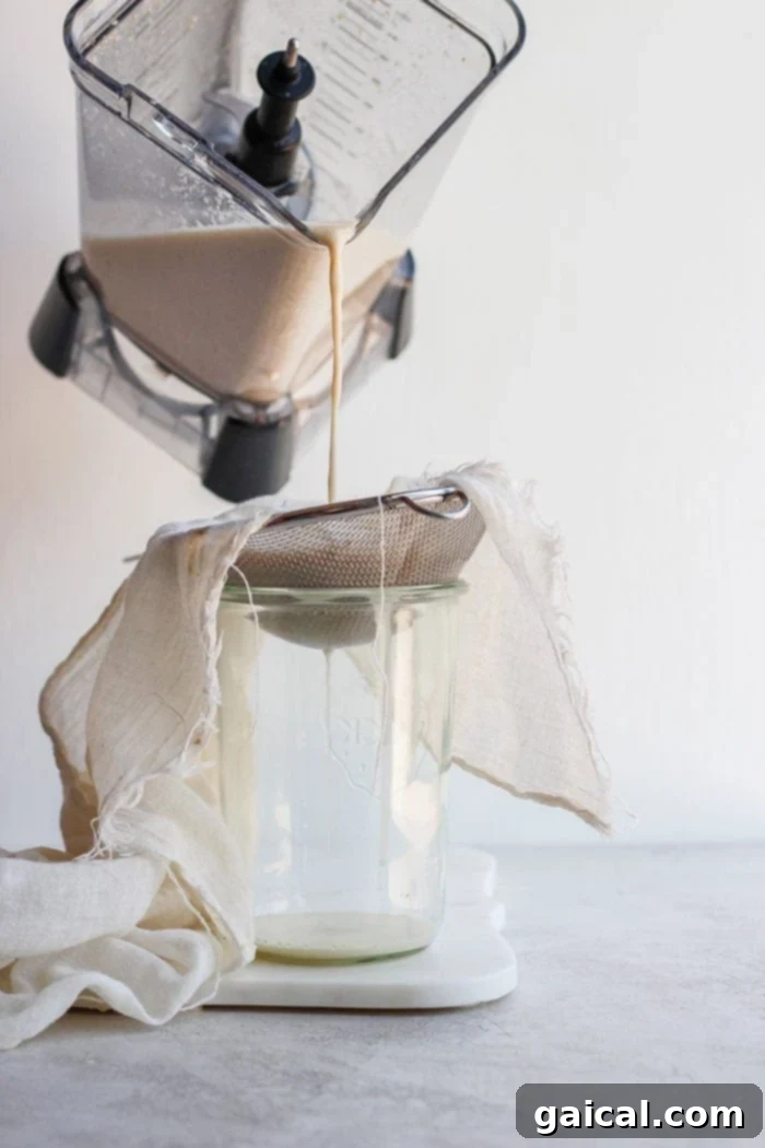Straining Homemade Oat Milk Homemade oat milk being carefully poured through a tea towel and fine-mesh sieve into a glass pitcher, illustrating the straining process.