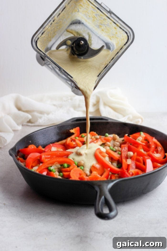 Rich and creamy cashew curry sauce being poured over sautéed vegetables in a large skillet, ready to simmer.
