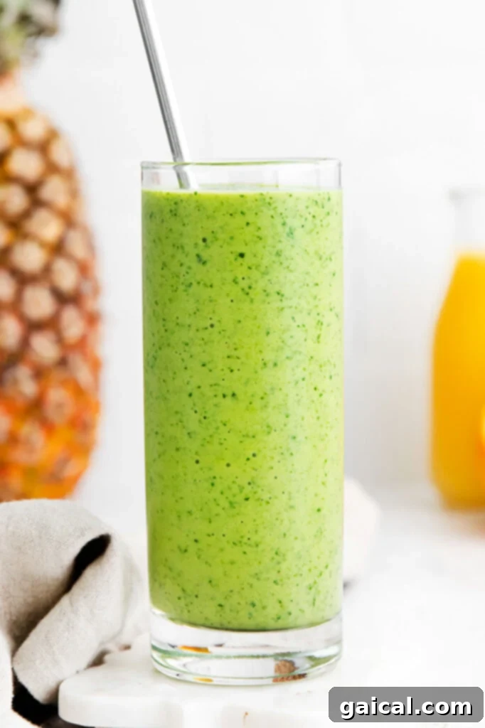 Island Green Power Smoothie 2 A vibrant green spinach smoothie in a tall glass, garnished with a pineapple wedge, next to fresh oranges and pineapples, set against a bright, natural background.