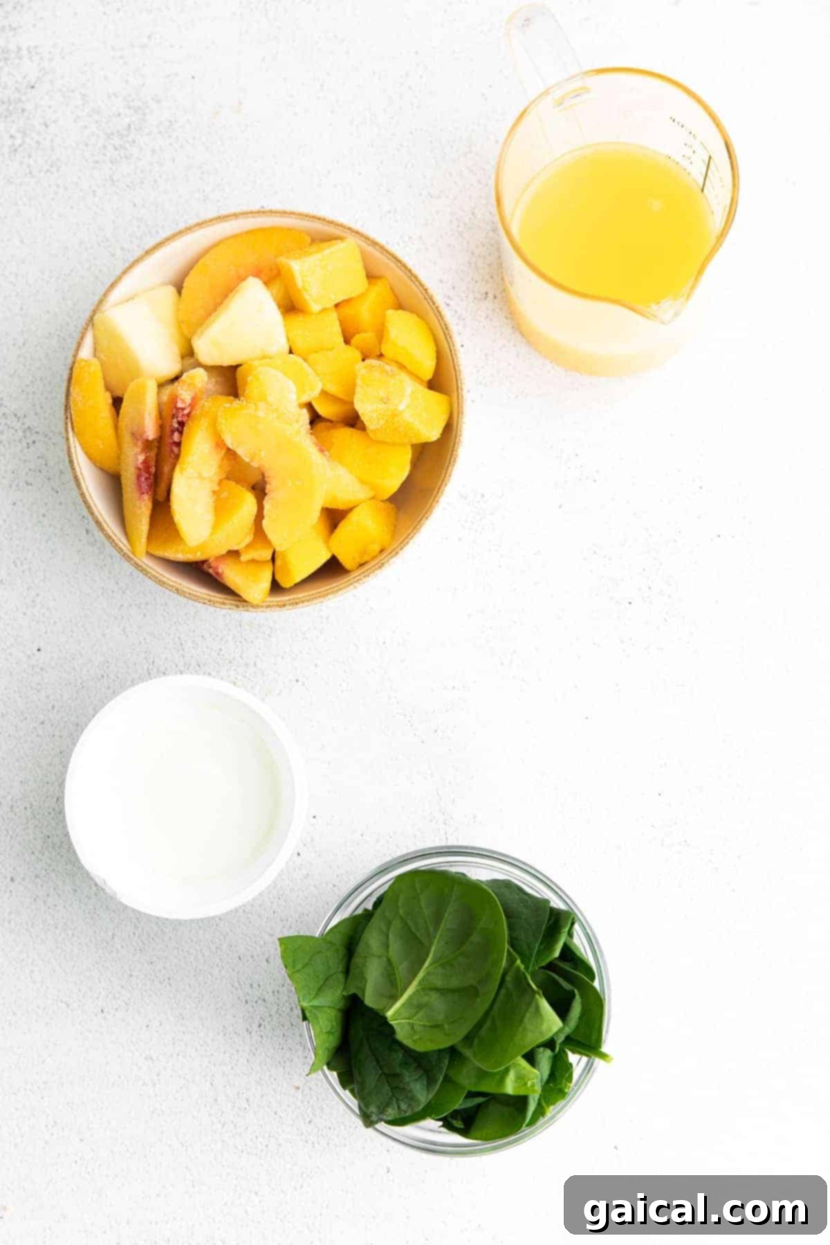 Island Green Power Smoothie 3 All the fresh and frozen ingredients for the tropical spinach smoothie, including a bag of frozen tropical fruit, fresh spinach leaves, Greek yogurt, and a glass of orange juice, neatly arranged in small bowls on a wooden surface.
