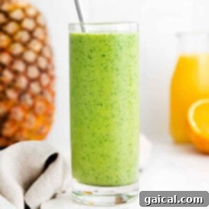 A vibrant green spinach smoothie in a glass with fresh oranges and pineapples in the background, signaling its tropical flavor profile.