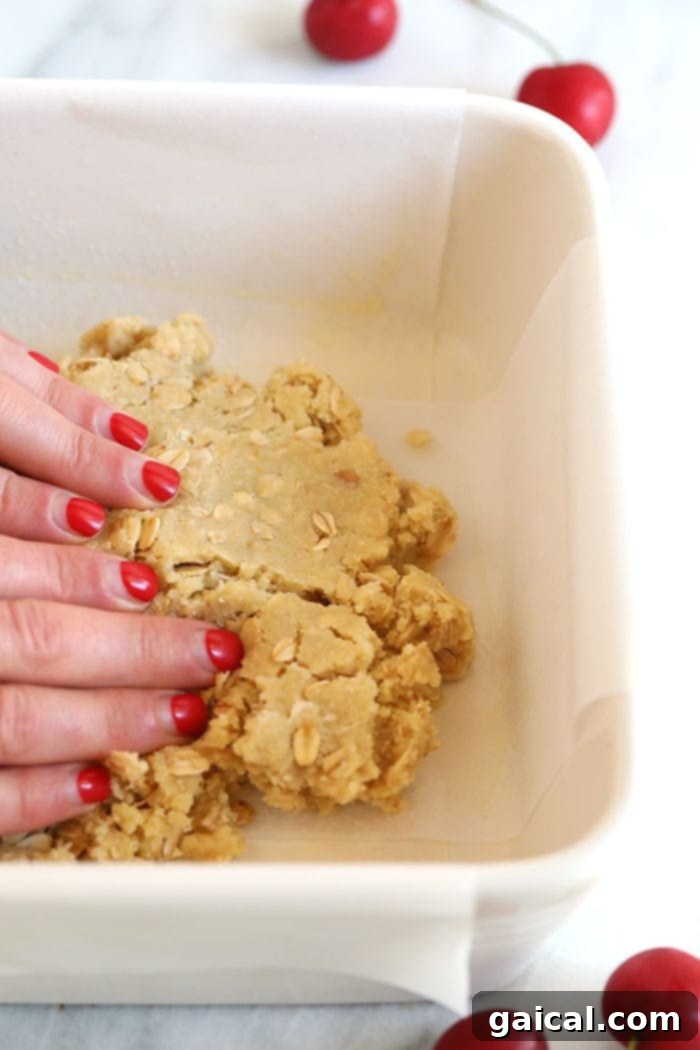 Ensure your crust is evenly pressed for the perfect base. Hands pressing almond flour and oat crust mixture into the bottom of a baking pan, creating a firm base for the cherry filling.