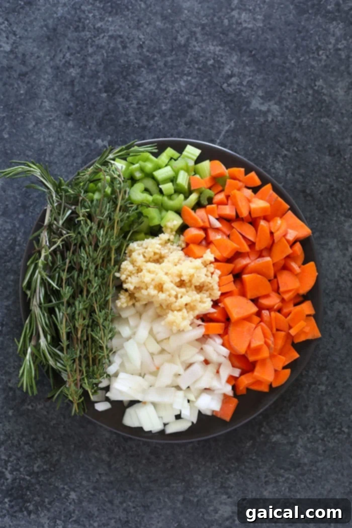 Fresh ingredients laid out on a white plate, including chicken breast, carrots, celery, onion, garlic, and fresh herbs.