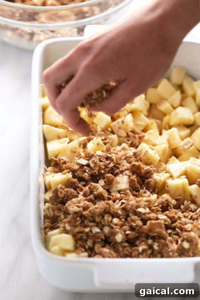 This delicious apple crisp recipe is made with a simple apple filling and a delicious crumble topping. Make this healthier, fall-inspired dessert today! A pair of hands sprinkling a generous layer of oat crumble topping over a bed of sliced apples in a baking dish, illustrating the assembly process of apple crisp.