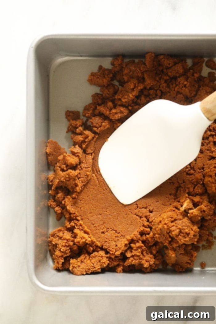 The crunchy gingersnap crust mixture being pressed firmly and evenly into a baking pan, preparing for the cheesecake filling.