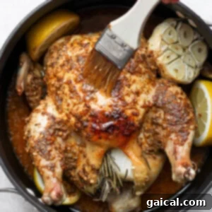 A person basting a whole roasted chicken with pan juices using a brush inside a hot oven.